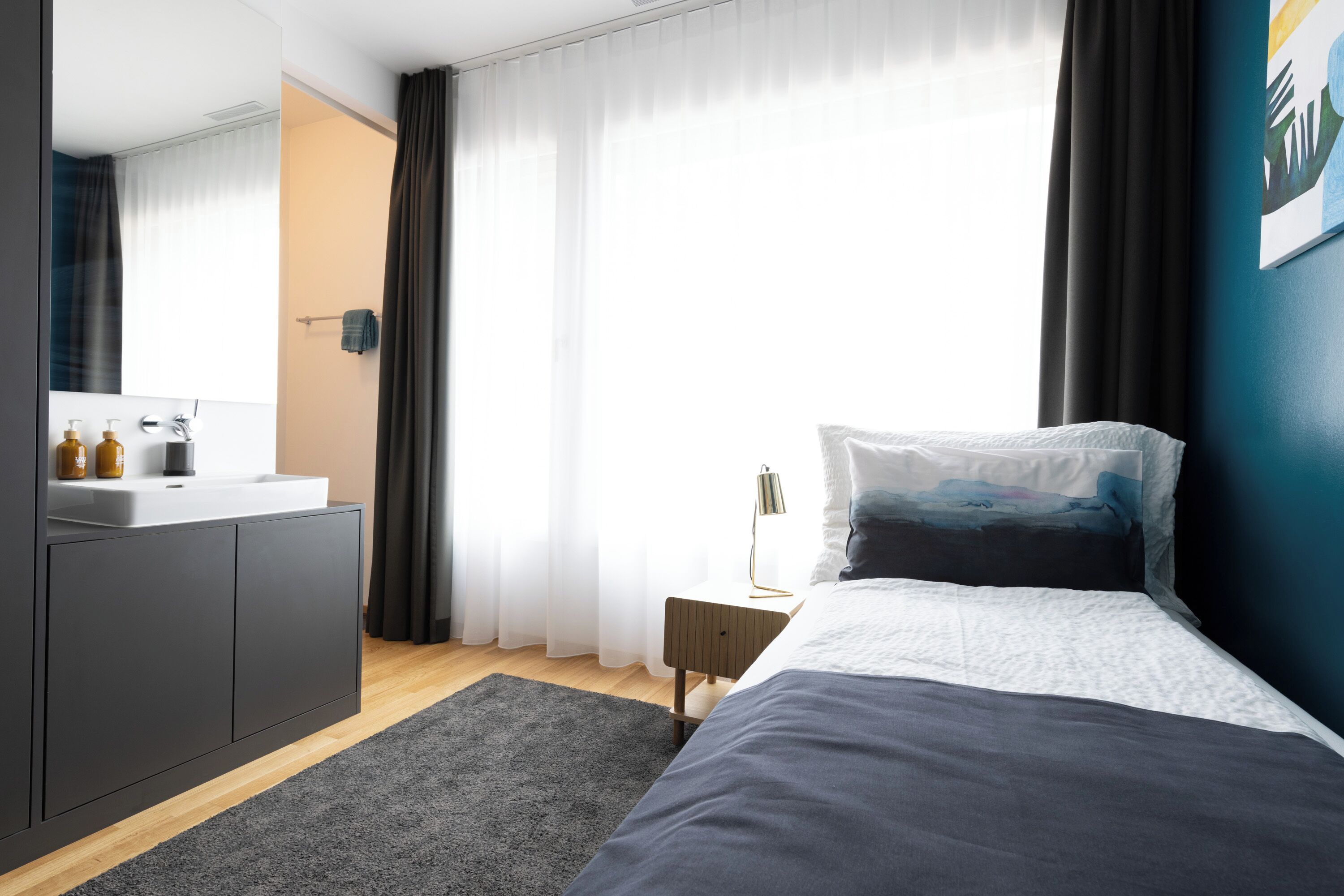 Una camera da letto moderna con un letto grande, un comodino, uno specchio, un bagno con un lavandino e una finestra con tende.