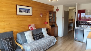 A cozy living area with a wooden wall, a sofa with patterned cushions, a small cabinet, and a kitchen area with a refrigerator and coffee machine. - Résidence Plein Sud, Front de Neige Centre, à Côté Télécabine 3400m (Les Deux Alpes)