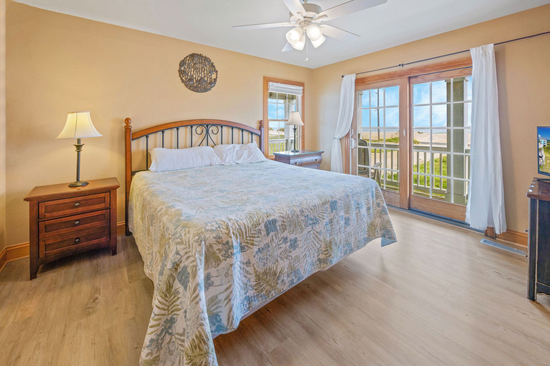 A bedroom with a bed, a nightstand, a ceiling fan, a window with a view of the beach, and a balcony with a chair.