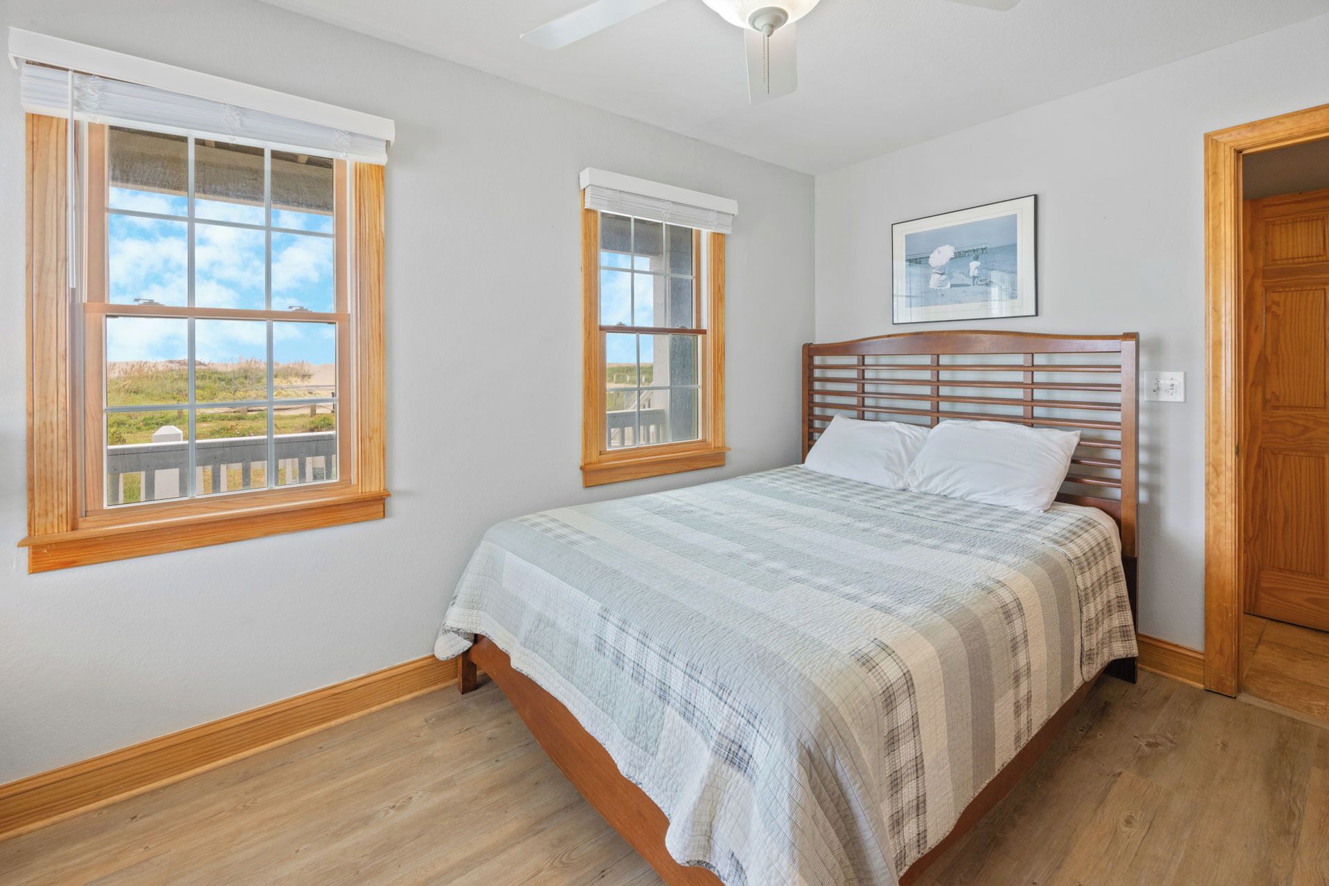 A bedroom with a wooden bed, two windows with blinds, a ceiling fan, and a framed picture on the wall.