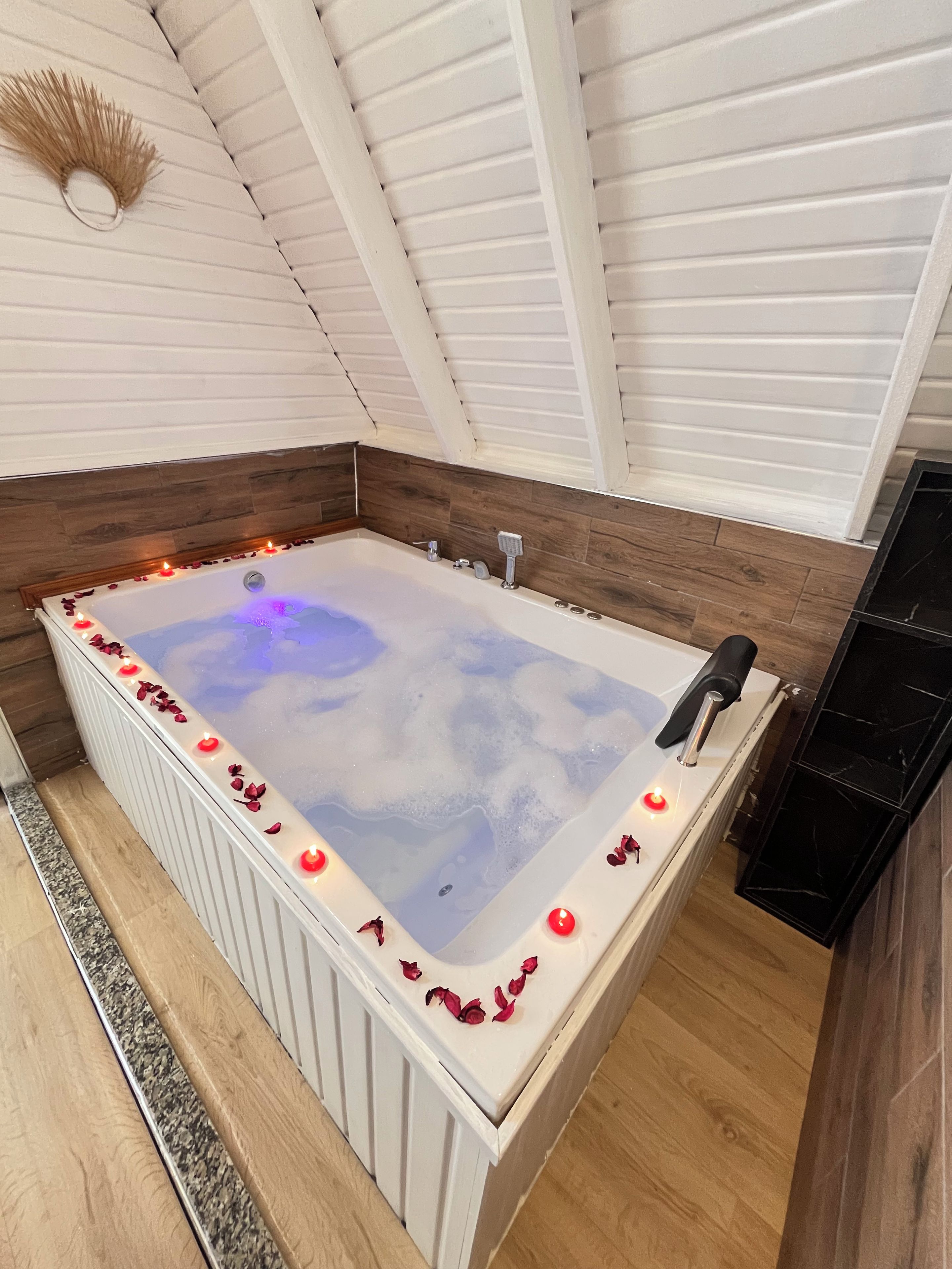 A rectangular hot tub with a wooden floor surrounding it, a ceiling with white beams, and a wall with wooden paneling.