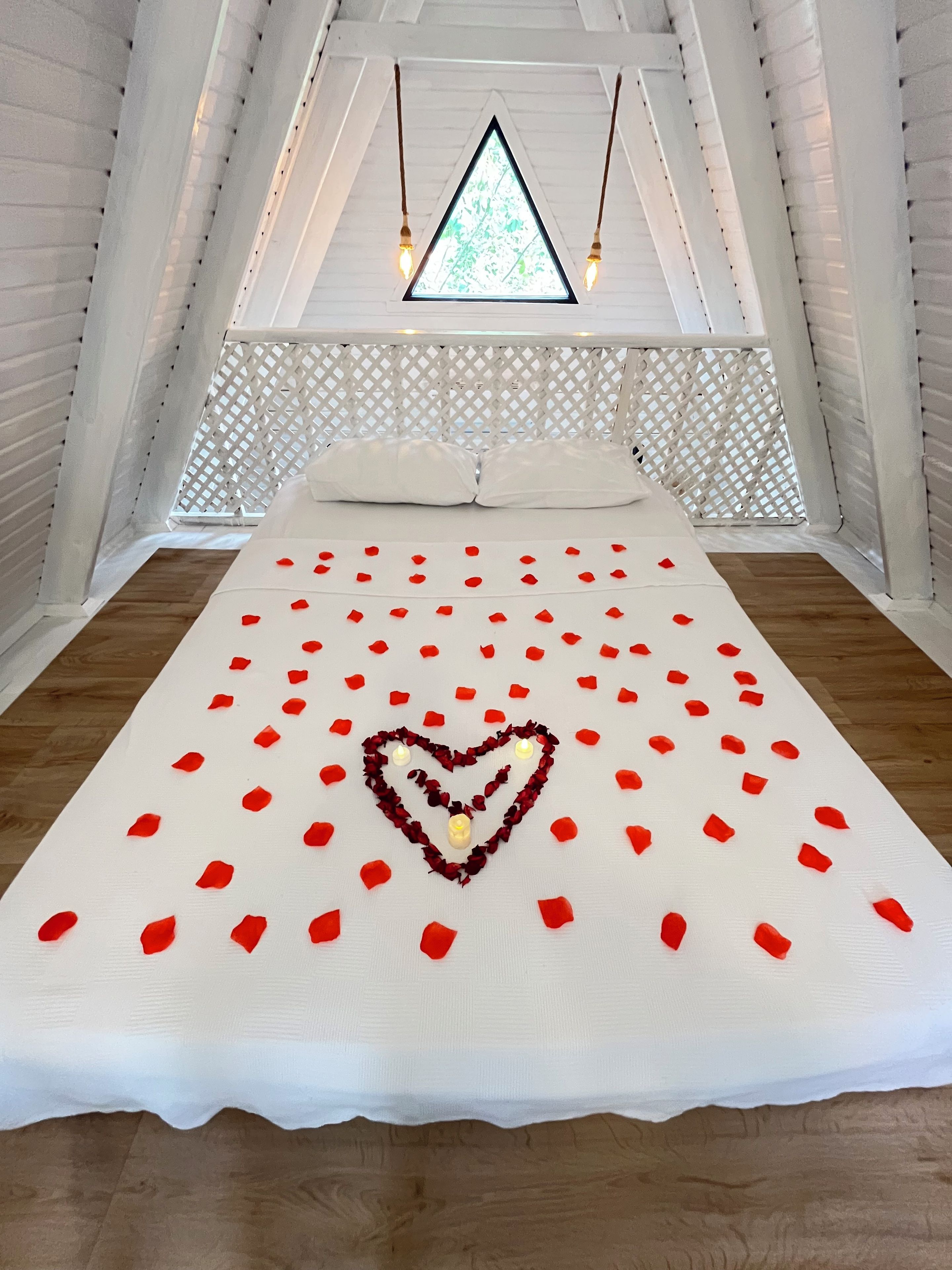 A bedroom with a bed, white bedding, a heart-shaped decoration made of rose petals, and a window with a view.