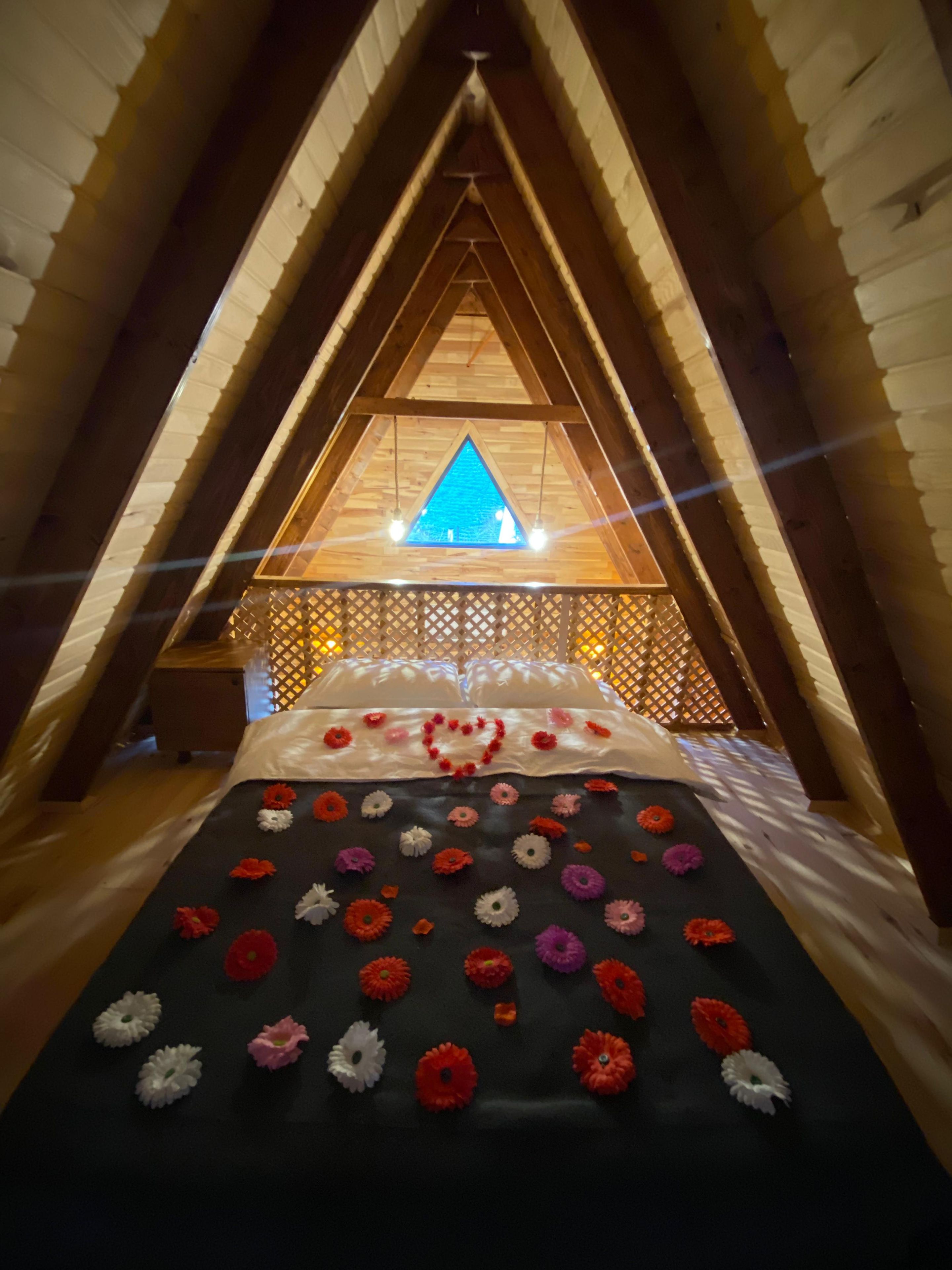 Una habitación en el ático con techo inclinado, una cama con un arreglo floral y un tragaluz triangular.