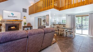 Een ruime woonkamer met een open haard, een grote hoekbank, een eettafel met stoelen, en een houten balkon.