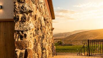 A stone building with a wooden roof and a paved walkway leading to a fenced area with a view of hills.