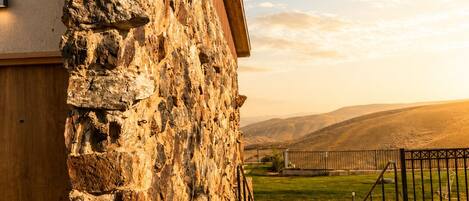 A stone building with a wooden roof and a paved walkway leading to a fenced area with a view of hills.