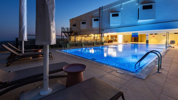 Una piscina con sillas reclinables y sombrillas, un área de estar techada y un edificio con varias ventanas y un letrero.