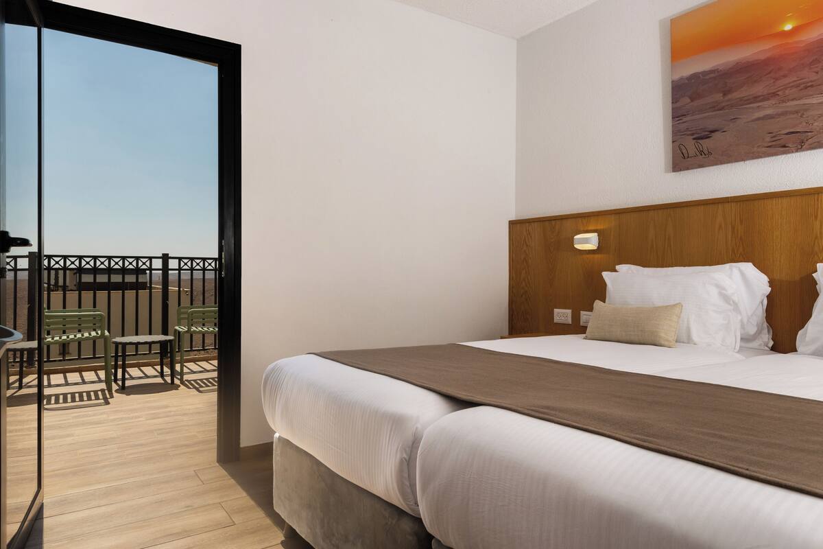 Habitación de hotel con cama, cabecero de madera, balcón con sillas y un cuadro en la pared.