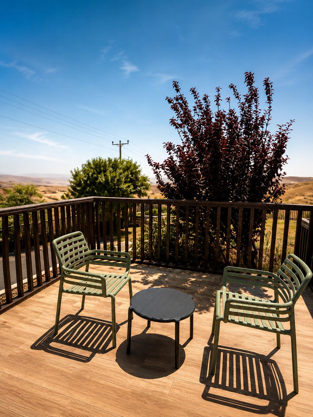 A patio with two green metal chairs and a small round table, surrounded by a wooden fence with a view of a hilly landscape.