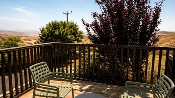 A patio with two green metal chairs and a small round table, surrounded by a wooden fence with a view of a hilly landscape.