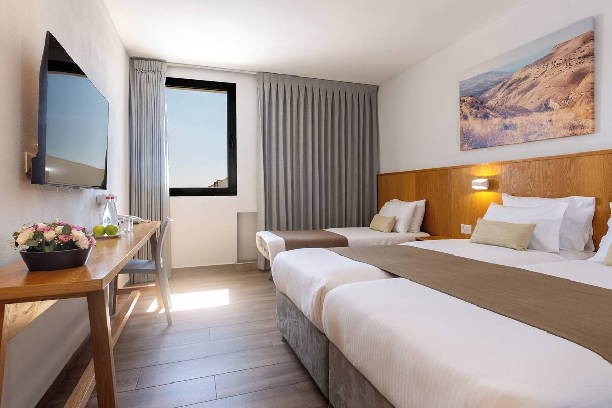 Habitación de hotel con cama, escritorio con televisor, ventana con cortinas y un cuadro en la pared.