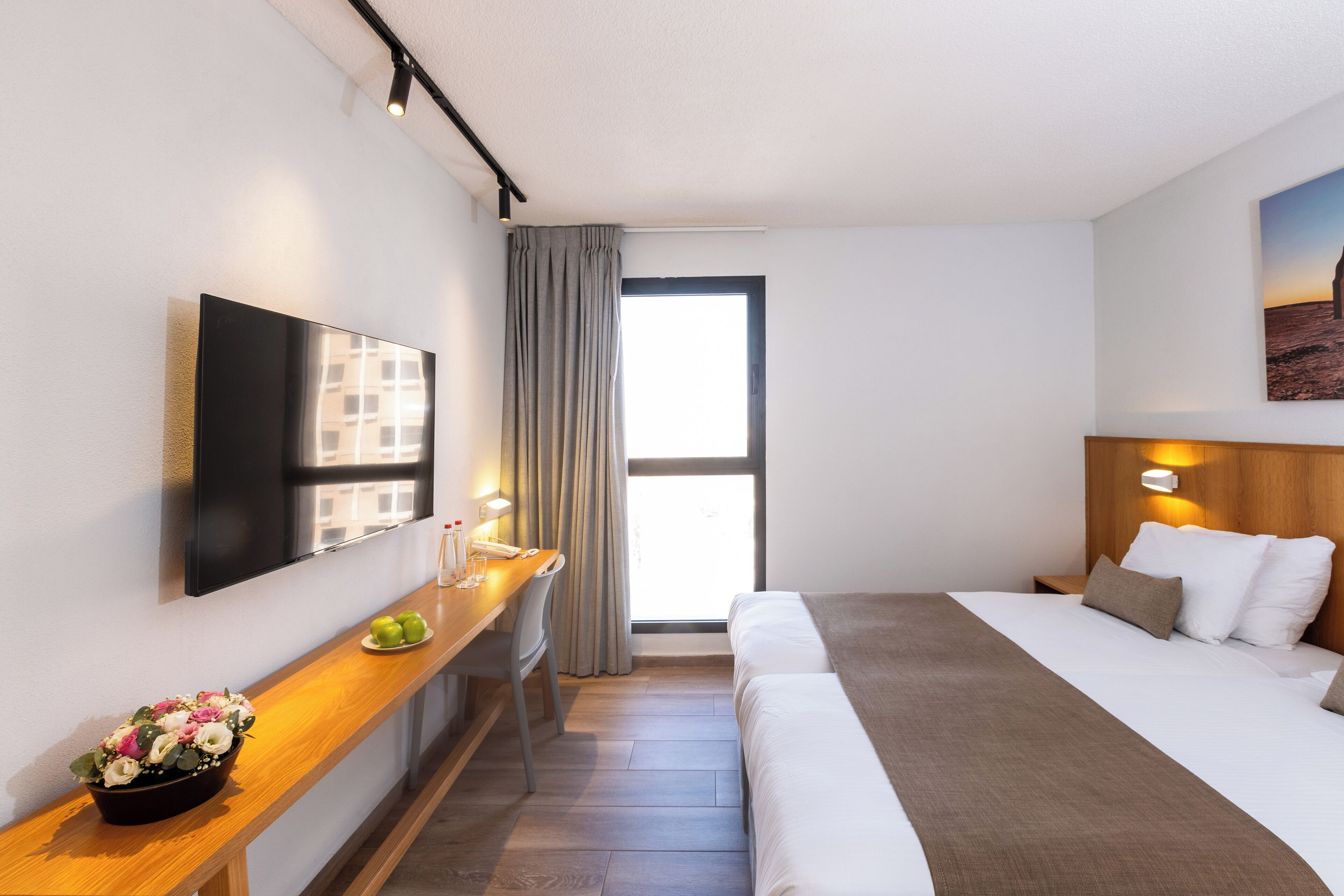 Una camera d'albergo moderna con schermo piatto, una scrivania in legno con lampada, un letto con lenzuola bianche e un'ampia finestra con tende.