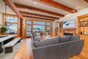 A cozy living room with a grey sofa, wooden beams, a fireplace, and a large window overlooking a snowy landscape.