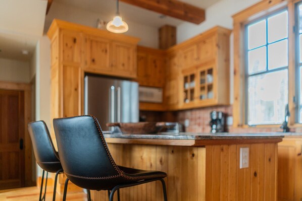 A kitchen with wooden cabinets, a stainless steel refrigerator, a countertop with a coffee maker, and two black chairs.
