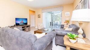 En moderne stue med en grå sofa, et fladskærms-tv, et spiseområde med et glasbord, og udsigt til det fri.