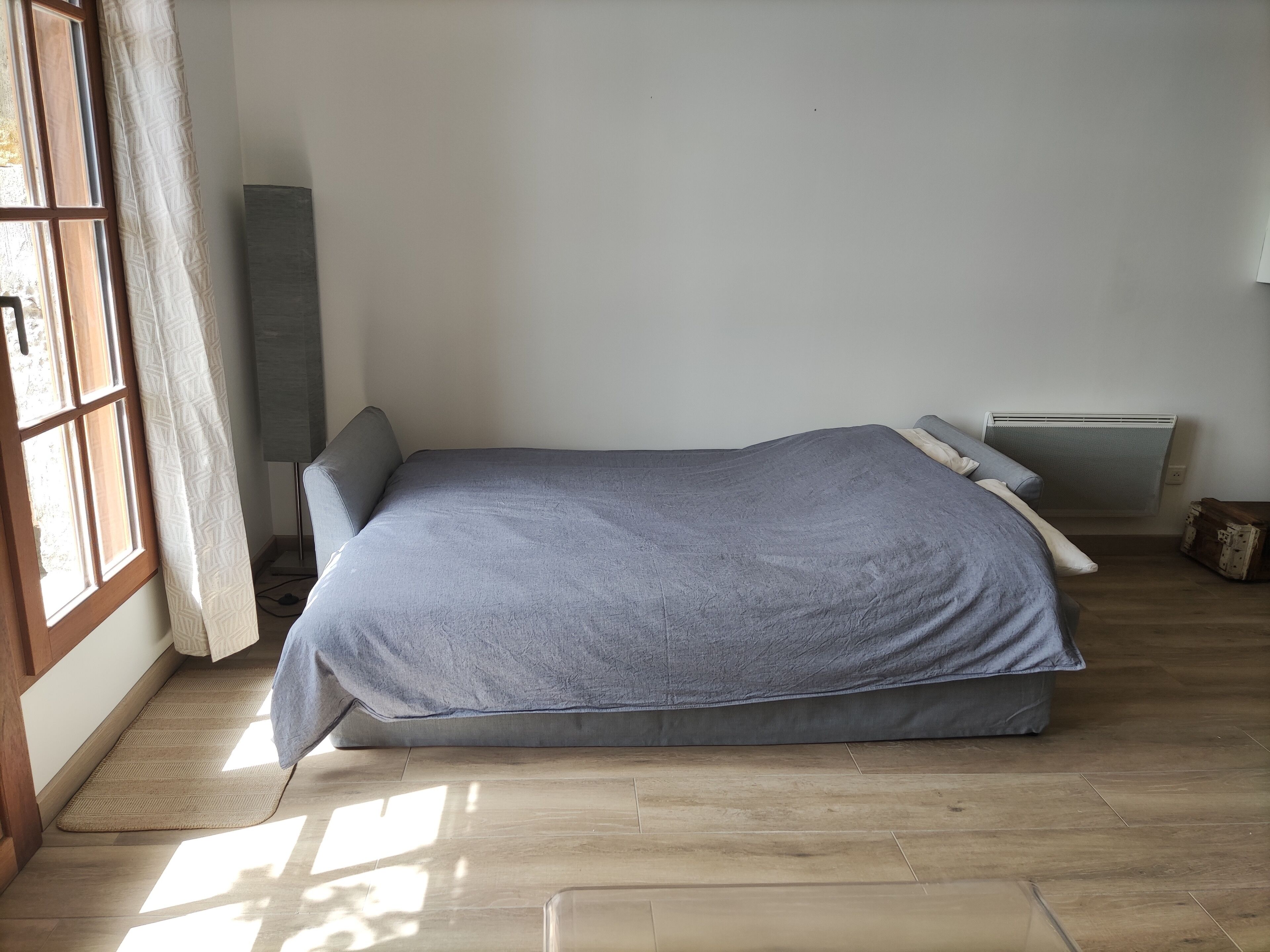 Un lit simple avec un matelas et des oreillers gris, un plancher en bois, une fenêtre avec des rideaux à motifs et un radiateur.