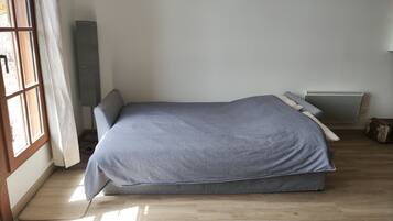 Un lit simple avec un matelas et des oreillers gris, un plancher en bois, une fenêtre avec des rideaux à motifs et un radiateur.