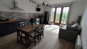 A modern kitchen with dark cabinets, a wooden dining table, and a large window. - Gîte de L'atelier (Saint-Privat)