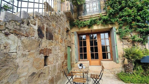 A courtyard with a stone wall, wooden furniture, and a balcony with greenery. - Gîte de L'atelier (Saint-Privat)