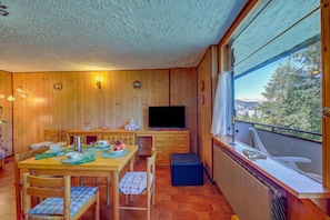 Quarto com painéis de madeira, uma mesa de jantar, cadeiras, uma televisão e uma janela com vista para árvores e montanhas.