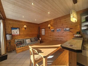 Interior acolhedor de uma cabana de madeira, com uma mesa de madeira, um sofá xadrez e uma área de cozinha com pia e armários.