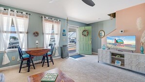 A living room with a dining table, chairs, a television, and a green door. - The Mermaid Lounge by Seaside Vacations & Sales (Chincoteague Island)