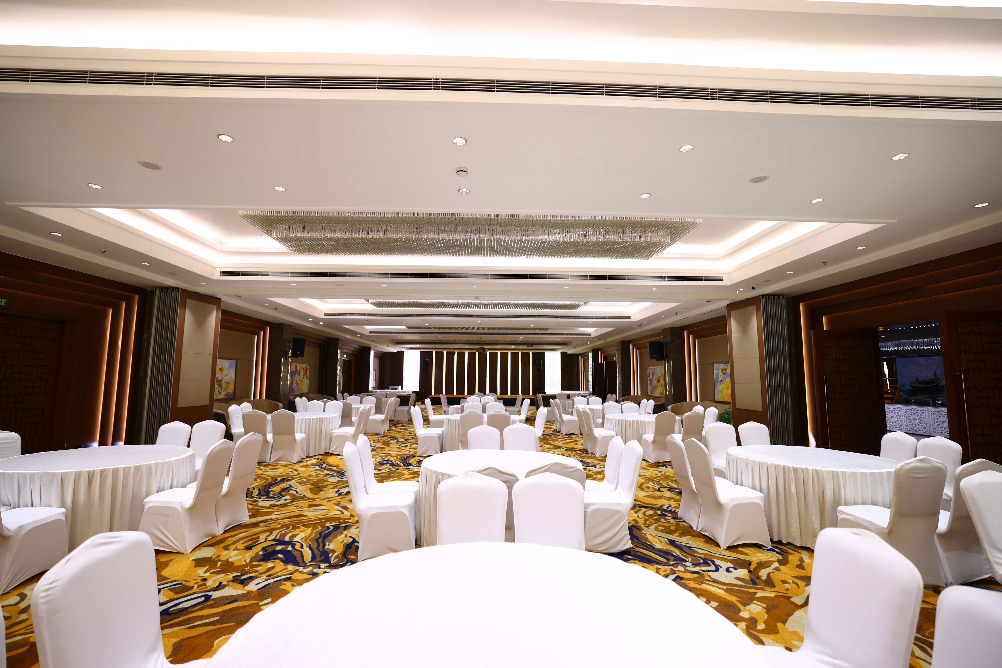 A large conference room with rows of white chairs and tables, a patterned carpet, and a modern chandelier.