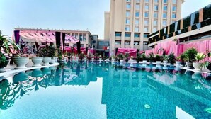 A hotel pool area with a clear pool, potted plants, and a building with pink curtains. - Mars Resorts (Meerut)