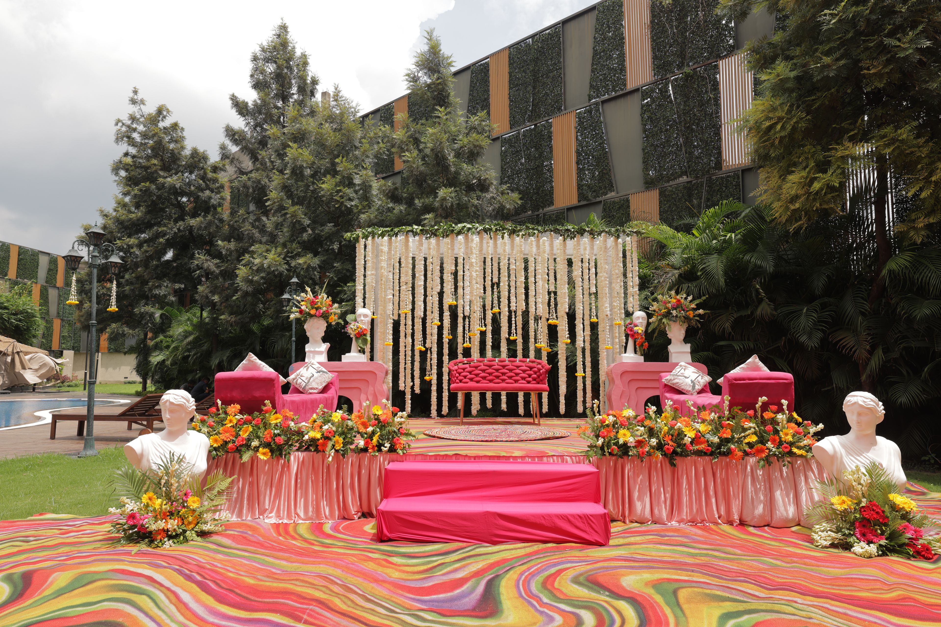Escenario para un evento al aire libre con un sofá rojo central, arreglos florales y esculturas de bustos clásicos.