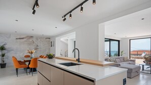 Eine moderne Küche mit einer zentralen Kochinsel, ein Essbereich mit orangefarbenen Stühlen und ein Wohnbereich mit einer Couch und Blick ins Freie.