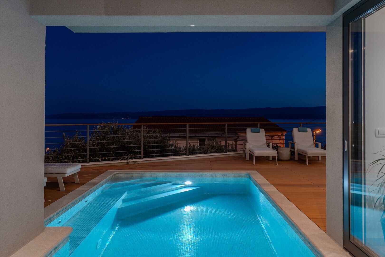 A rooftop pool with a view of the sea at night, surrounded by lounge chairs and a wooden deck.