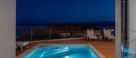 A rooftop pool with a view of the sea at night, surrounded by lounge chairs and a wooden deck.