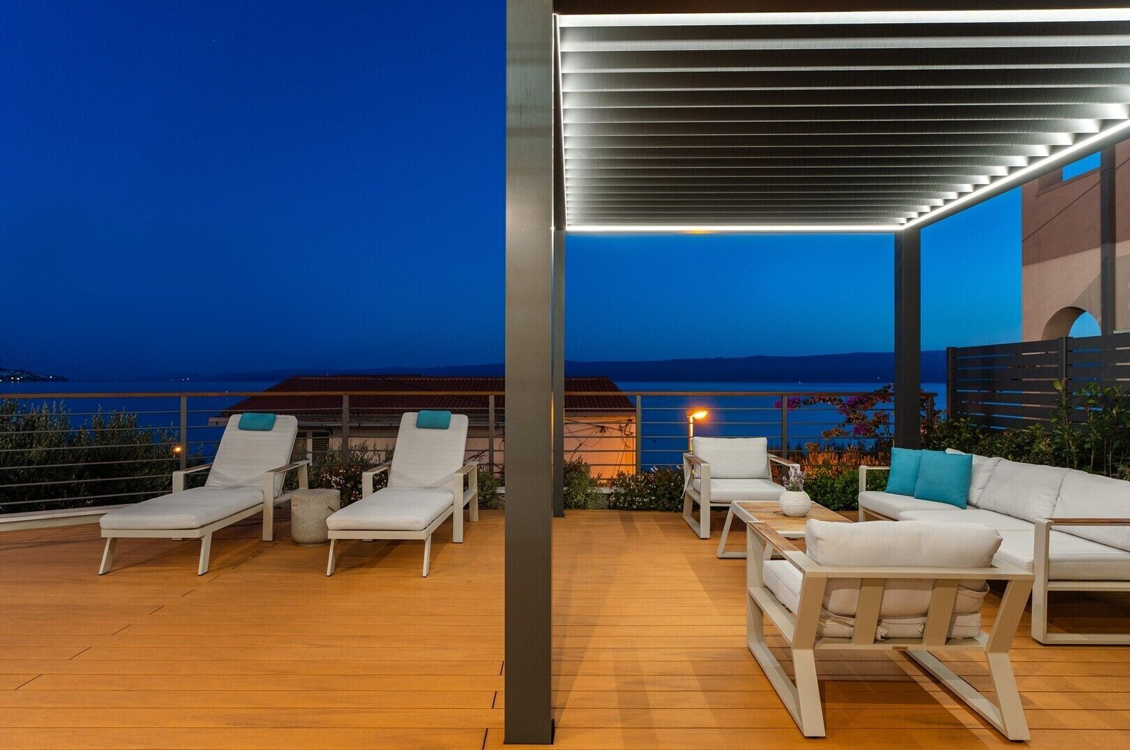 Eine Dachterrasse mit weißen Liegestühlen und einem Holztisch, mit Blick auf ein Gewässer bei Dämmerung.