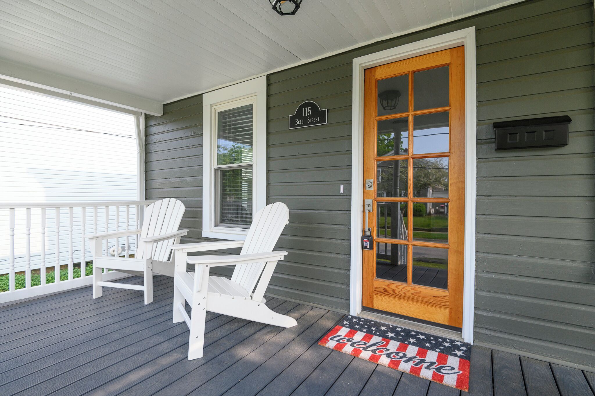 A porch with two white Adirondack chairs, a wooden door with a glass panel, and a welcome mat with stars and stripes.