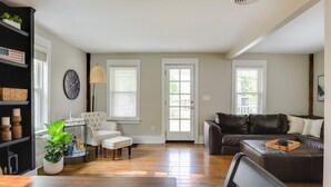 Sala de estar con pisos de madera, un sofá de cuero oscuro, un sillón blanco y una mesa de centro. Se observa una estantería con adornos y una planta en maceta.