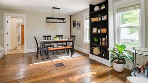 Un comedor con mesa y sillas de madera, una lámpara colgante y una estantería negra con objetos decorativos.