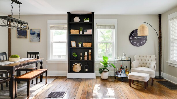 Un comedor con una mesa de madera y bancos, una estantería negra con objetos decorativos, un sillón blanco y un reloj de pared redondo.