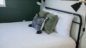 Uma cama com colchão branco, estrutura de metal preto e duas almofadas, uma verde e outra estampada.