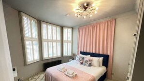 Ein Schlafzimmer mit einem Bett, einem Heizkörper, einem Fenster mit Jalousien, einer Deckenleuchte und einem pinkfarbenen Vorhang.