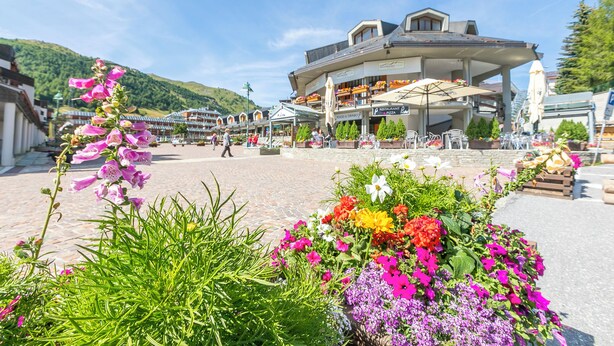 Ein Hotel mit überdachter Außenbestuhlung, Blumenarrangements und einer Bergkulisse.