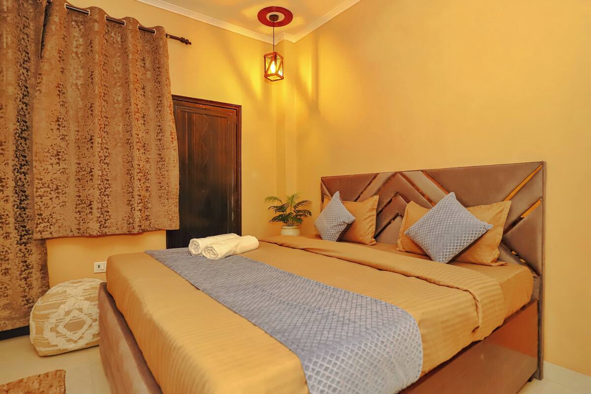 A double bed with a wooden headboard, a bedside table with a plant, and a curtain with a floral pattern.