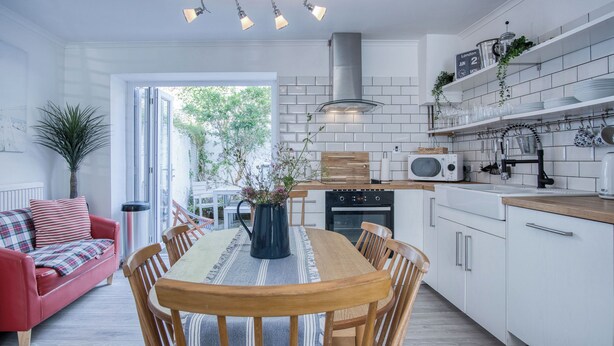 A modern kitchen with white cabinets, a wooden dining table, and a red sofa in the living area. - Norton Cottage - 3 Bedroom Cottage - Tenby (Tenby)