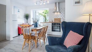 A modern kitchen with a built-in oven, a dining area with wooden chairs, and a blue armchair with a striped pillow. - Norton Cottage - 3 Bedroom Cottage - Tenby (Tenby)