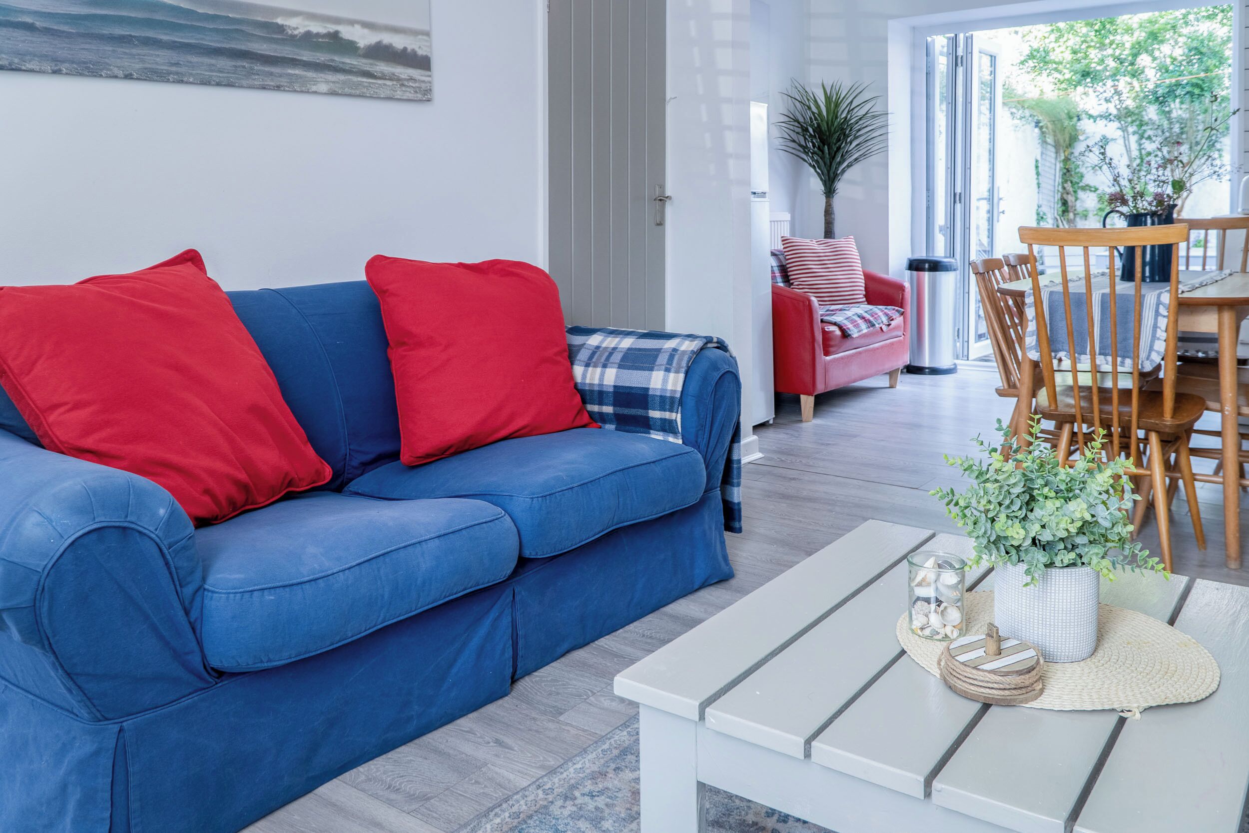 A modern living room with a blue sofa, red cushions, a white coffee table, and a dining area with wooden chairs and a table.
