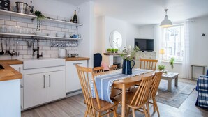A modern kitchen with white cabinets, wooden chairs, and a dining table. There is a TV and a window with curtains. - Norton Cottage - 3 Bedroom Cottage - Tenby (Tenby)