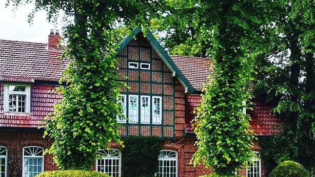 A traditional brick building with a green roof and white windows, surrounded by well-maintained hedges and trees. - Hotel Hof Kirchhorst (Gross Wittensee)