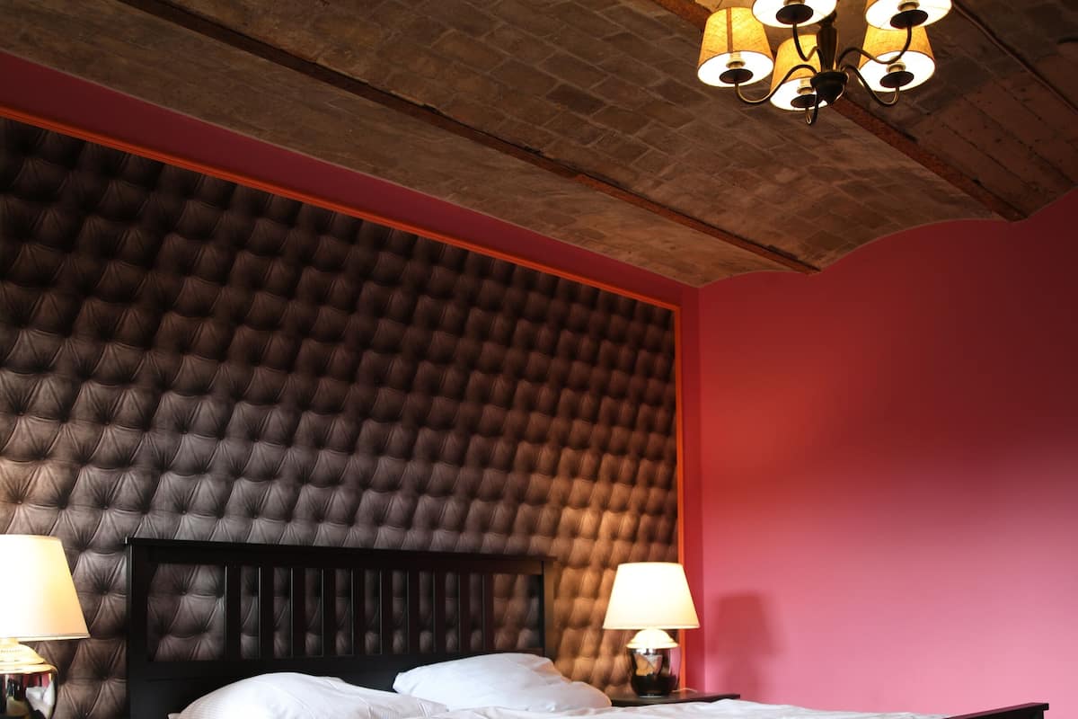 Une chambre à coucher avec une tête de lit capitonnée, un lit recouvert de draps blancs, deux lampes de chevet et un plafond en bois agrémenté d’un lustre.