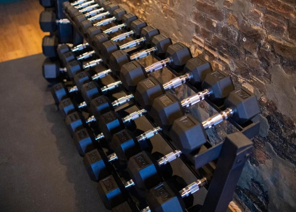 A row of black dumbbells with silver handles, arranged on a black mat against a brick wall.