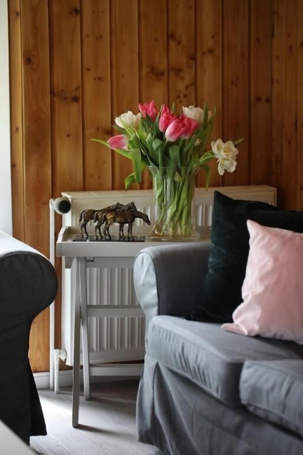 Una habitación con un sofá gris, un radiador blanco, un jarrón con flores y una mesita con una figurita de un caballo.