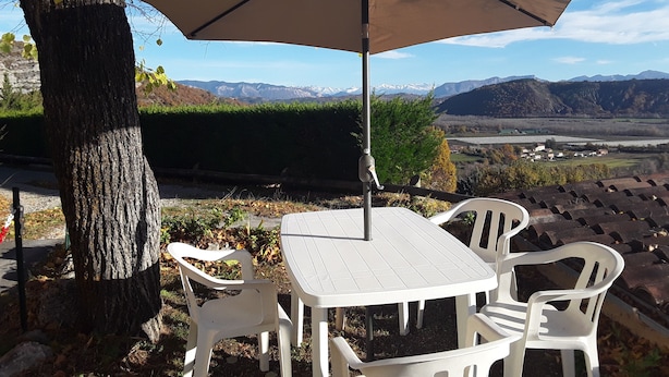Ein Tisch und Stühle unter einem Sonnenschirm, vor einer Bergkulisse und blauem Himmel.